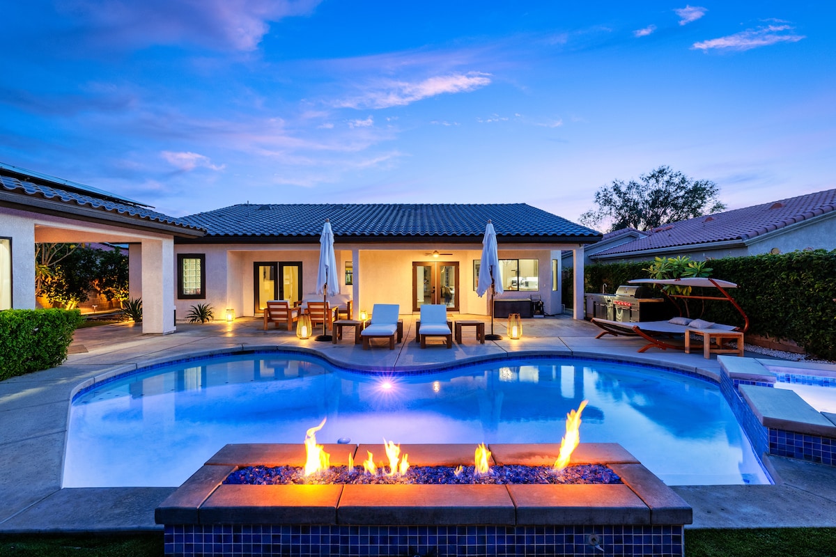 An inviting outdoor space features a shimmering pool with flames dancing atop a stone feature. Comfortable lounge chairs surround the pool, with umbrellas providing shade. The modern guesthouse stands in the background, softly illuminated by ambient lighting as the evening sky transitions to twilight.