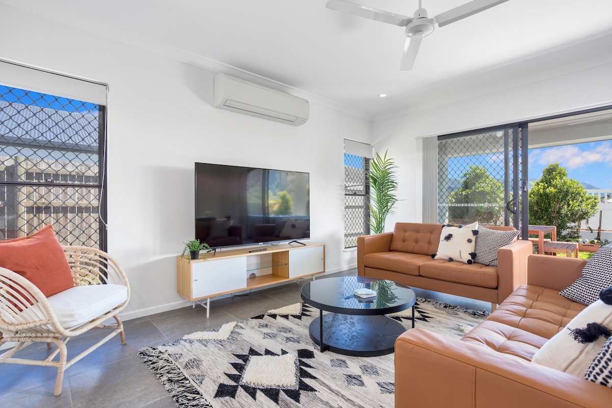 The open-plan living area highlights a comfortable seating arrangement, including a leather sofa and a rattan chair. A round coffee table sits in the center, while a large television is mounted on the wall, surrounded by large windows that provide natural light.
