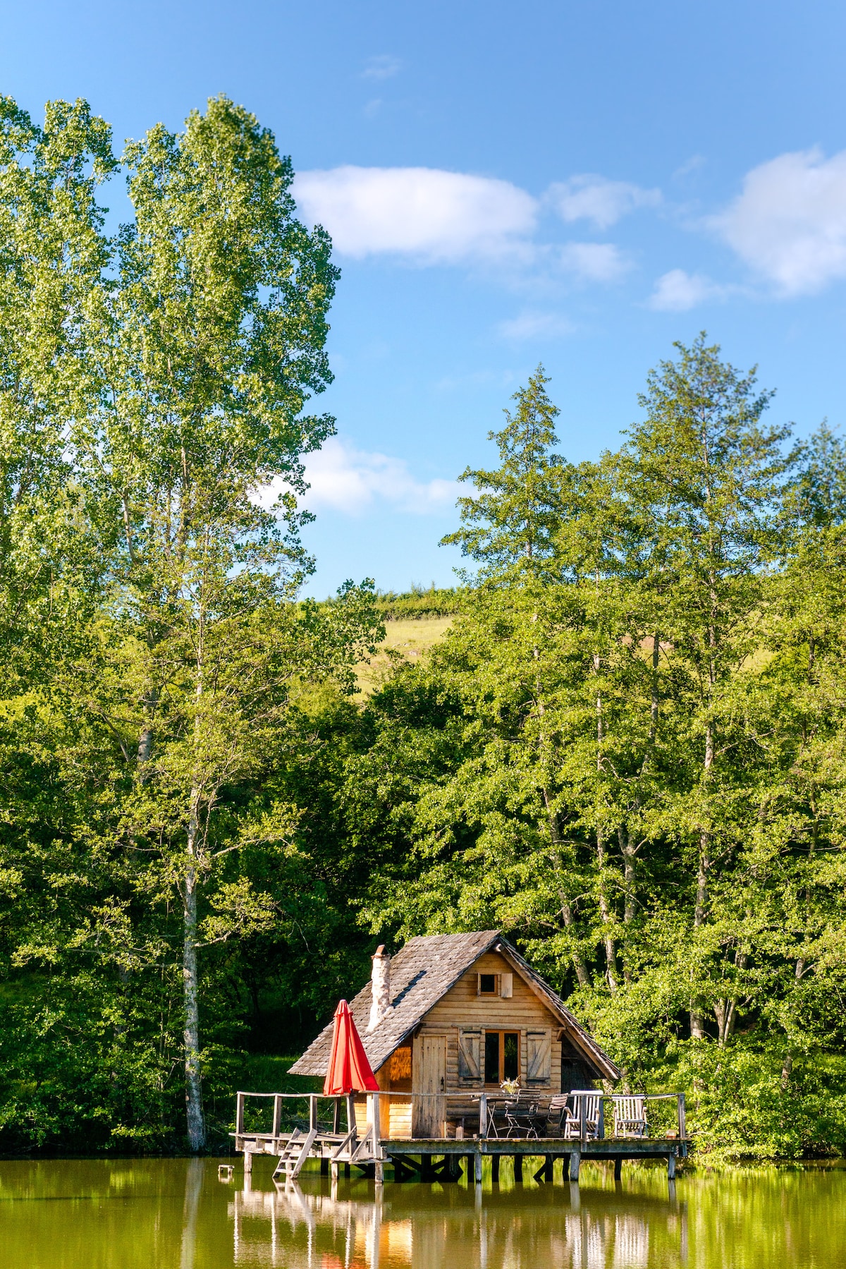 A charming wooden cabin is situated on stilts over a serene pond, surrounded by lush trees. A vibrant red umbrella is positioned on the terrace, providing a pop of color against the natural backdrop. The sky is bright with scattered clouds.