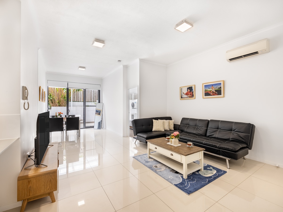 A spacious living area features a comfortable black sofa and a wooden coffee table, creating a relaxed atmosphere. A smart TV is positioned on a cabinet, and light from large sliding doors brightens the room. Artwork adorns the walls, complementing the modern decor.