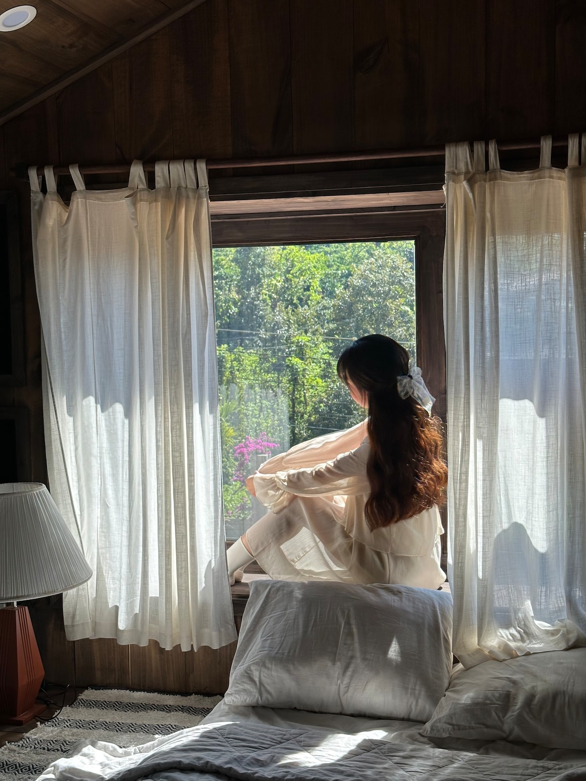 A serene indoor scene showcases a person seated on a windowsill, gazing out at the lush greenery. Soft natural light filters through sheer curtains, illuminating the room. A comfortable bed with white linens is positioned in the foreground, enhancing the peaceful ambiance.