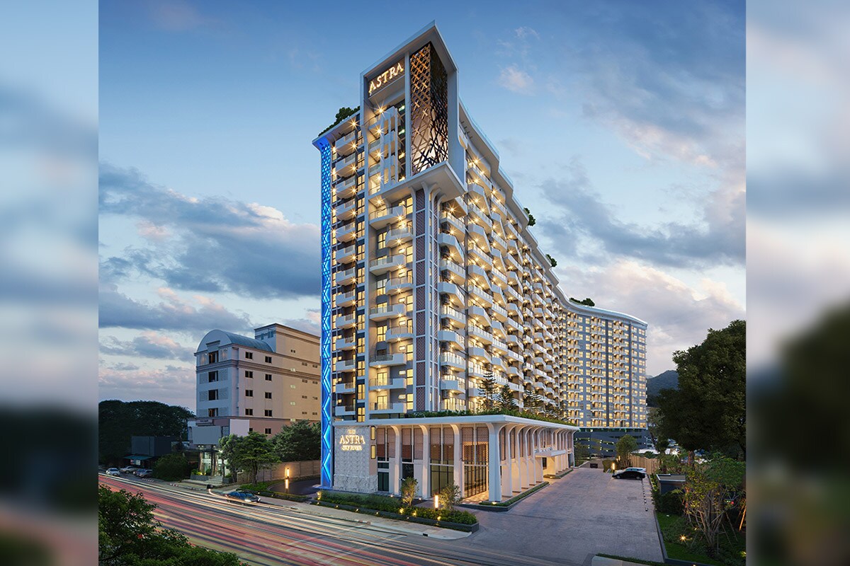 The modern condominium building features a striking architectural design with large balconies and illuminated facades. The entrance is marked by a curved overhang, while greenery is integrated throughout. The surroundings include other buildings and roads, enhancing the urban setting.