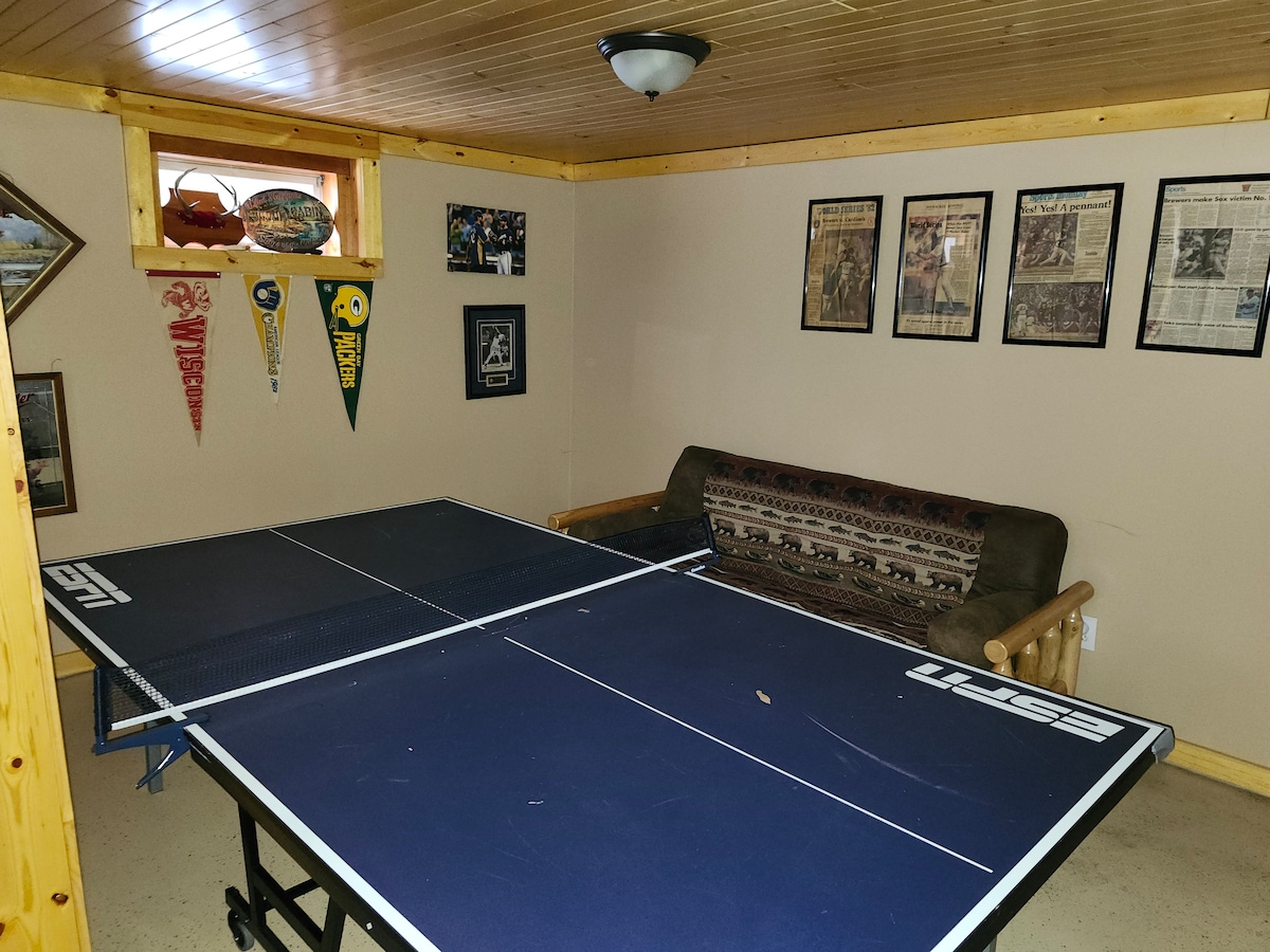 A game room is featured, showcasing a ping pong table positioned in the center. Surrounding walls are decorated with framed articles and memorabilia, including team pennants. A sofa is placed against one wall, providing a cozy spot to relax between games.