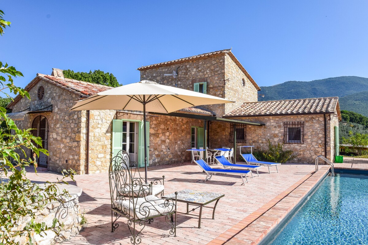 A charming villa made of natural stone displays a spacious pool area featuring sun loungers and a shaded umbrella. The facade is complemented by green shutters and large windows, with scenic hills visible in the background under a clear blue sky.