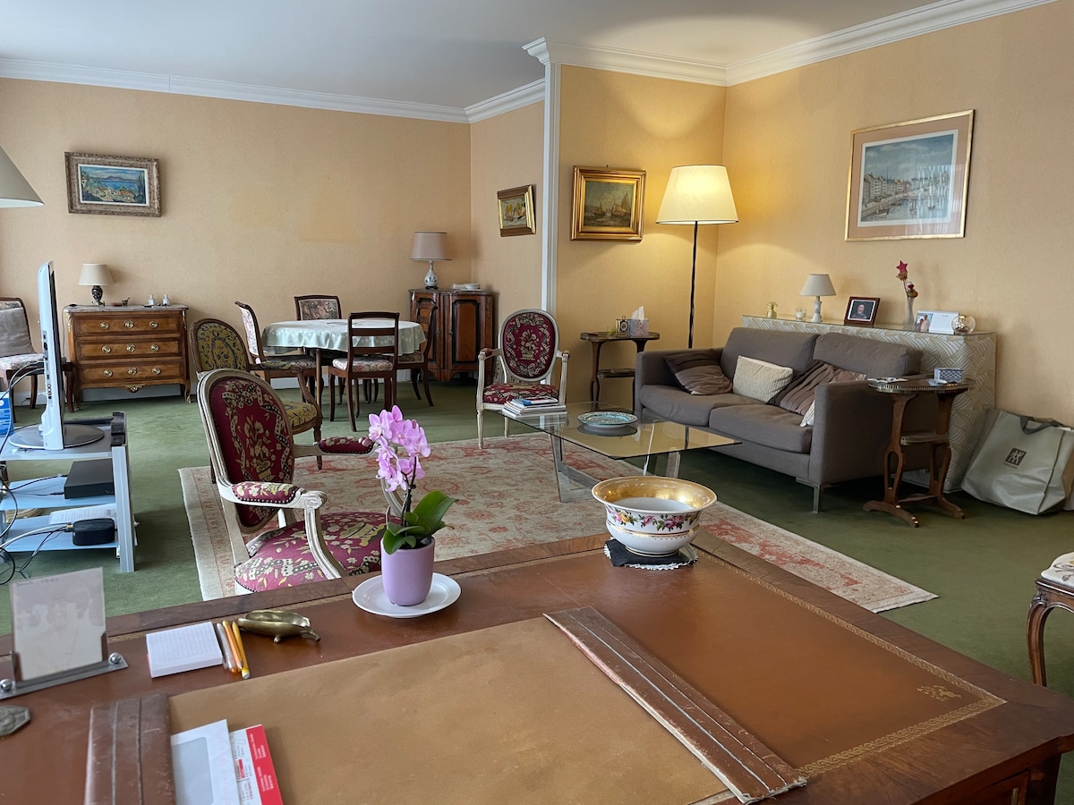 A bright common area is filled with various seating options, including a sofa and several chairs. A dining table is visible in the background. A decorative rug lies on the floor. Natural light enters through the windows, highlighting artworks on the walls.