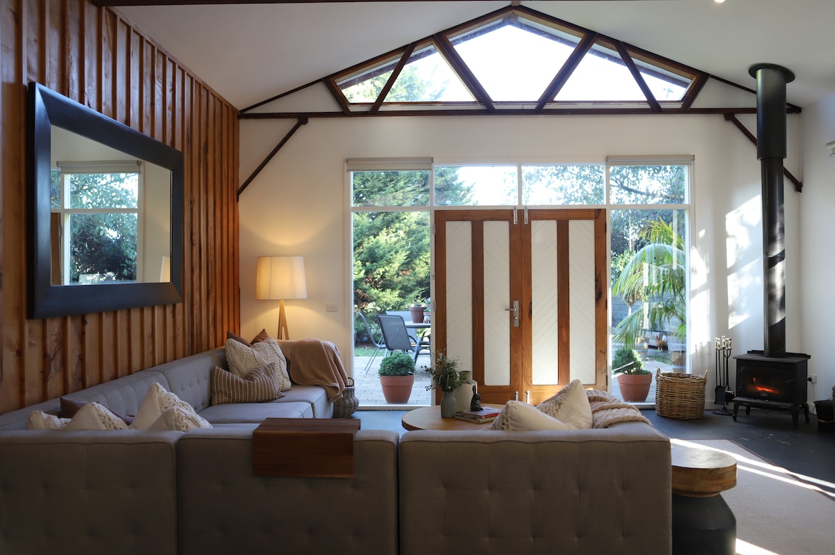 The inviting living area features a spacious sectional couch adorned with various cushions. Large windows allow natural light to fill the room, highlighting the wooden accents and elegant mirror. A wood-burning heater adds warmth, while a view of the courtyard greets guests through double doors.