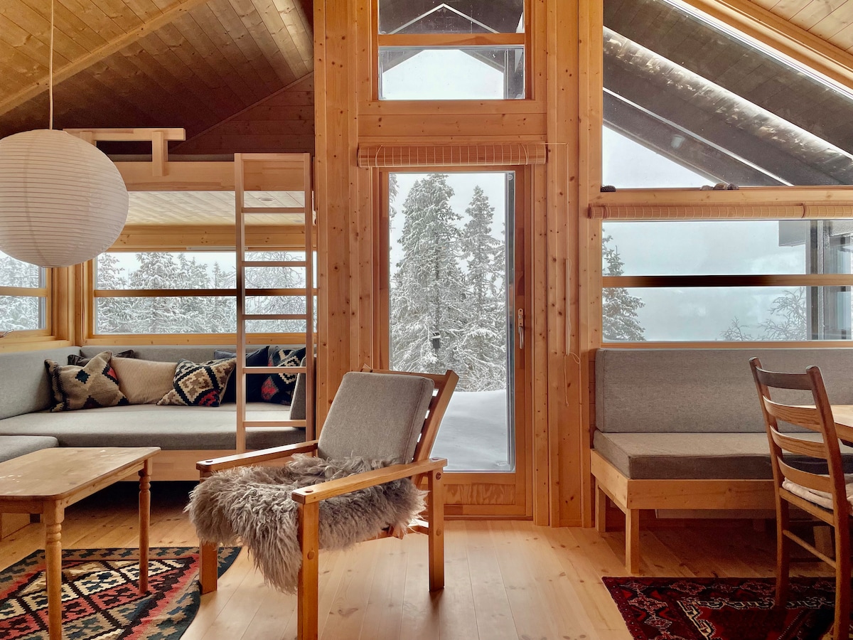 A bright living area is presented, featuring large windows that showcase snowy trees outside. Wooden walls and ceilings create a warm ambiance. Comfortable seating options include a plush sofa and a minimalist chair. A small wooden table and patterned rugs add character to the space.