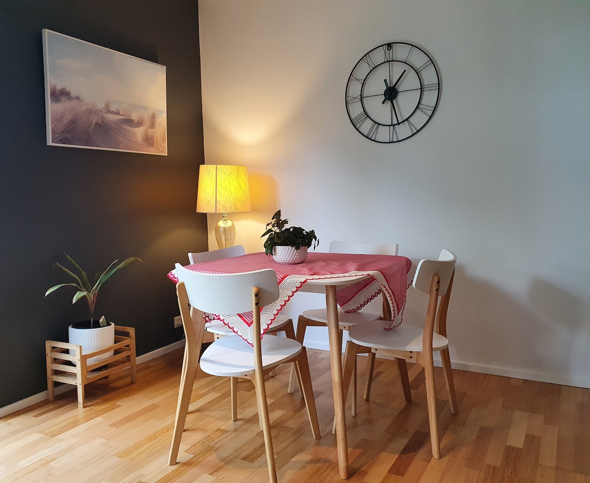 A dining area features a round table surrounded by six white chairs. A potted plant decorates the table, which is covered with a red and white table runner. A wall clock and a lamp add to the ambiance, highlighted against a contrasting dark wall.