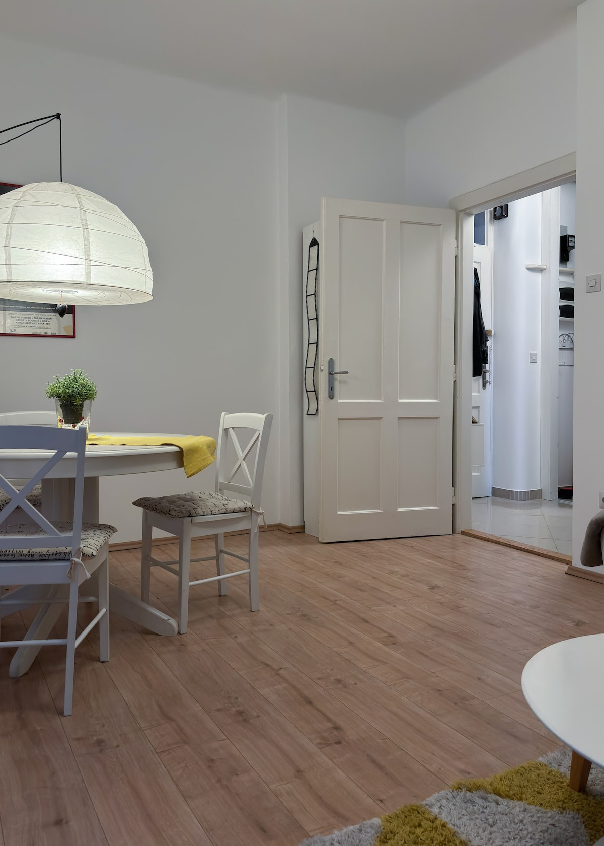 A bright dining area features a round table surrounded by two white chairs. A soft yellow tablecloth adds a pop of color. White walls create an open feel, and a doorway leads to an unseen space. A small potted plant rests on the table, enhancing the simplicity.