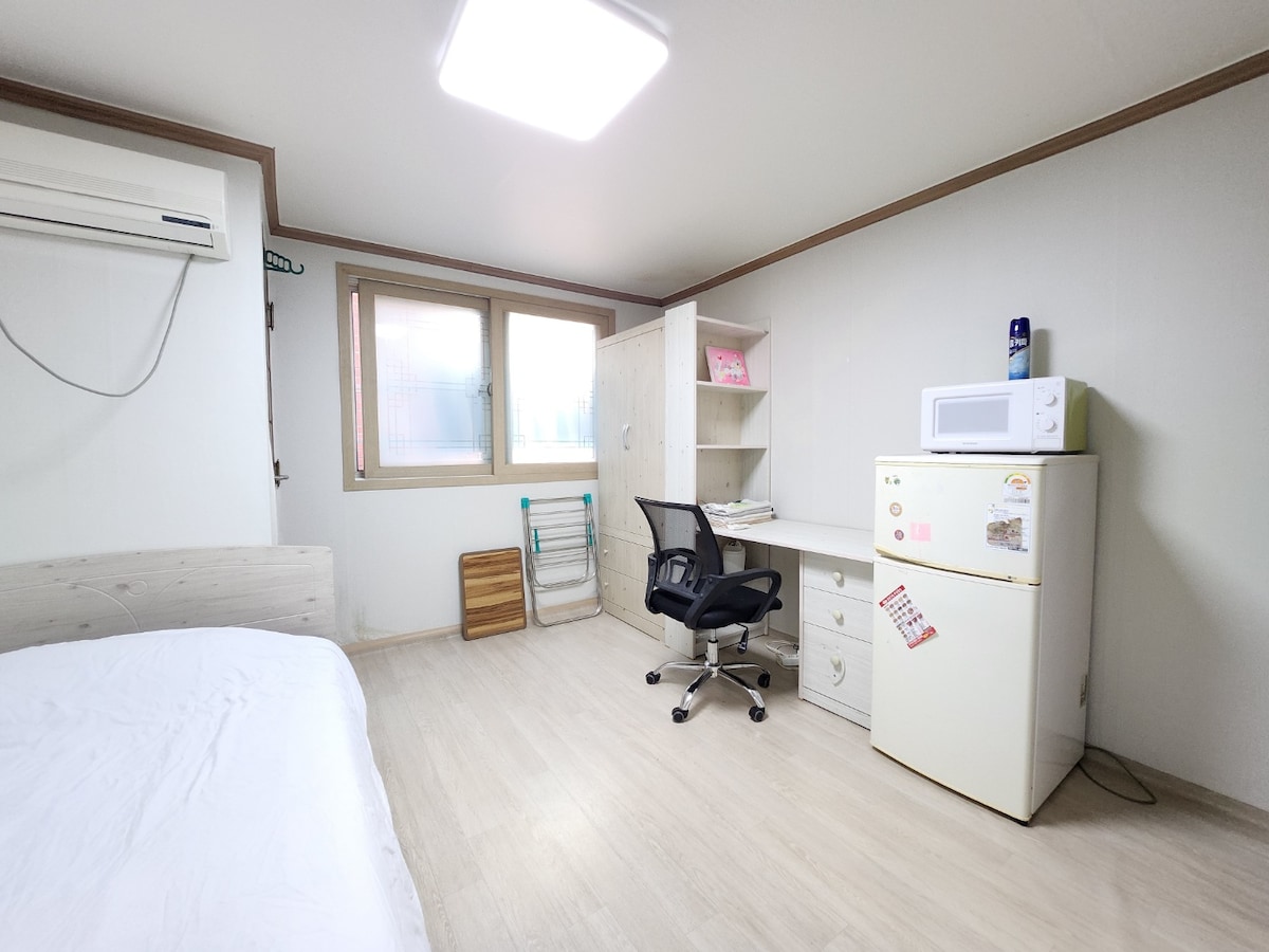 A clean and cozy room is featured, showcasing a spacious bed on one side. A desk with a chair and a microwave is positioned opposite a small refrigerator. Natural light enters through a window, while a clothes rack is present near the corner.