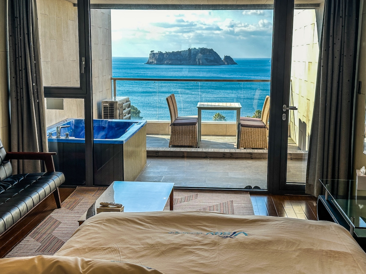 A spacious room offers a view of the ocean and a nearby island through large glass doors. An outdoor terrace features a dining table and chairs, while a heated bath is situated close to the bed. Natural light illuminates the area.