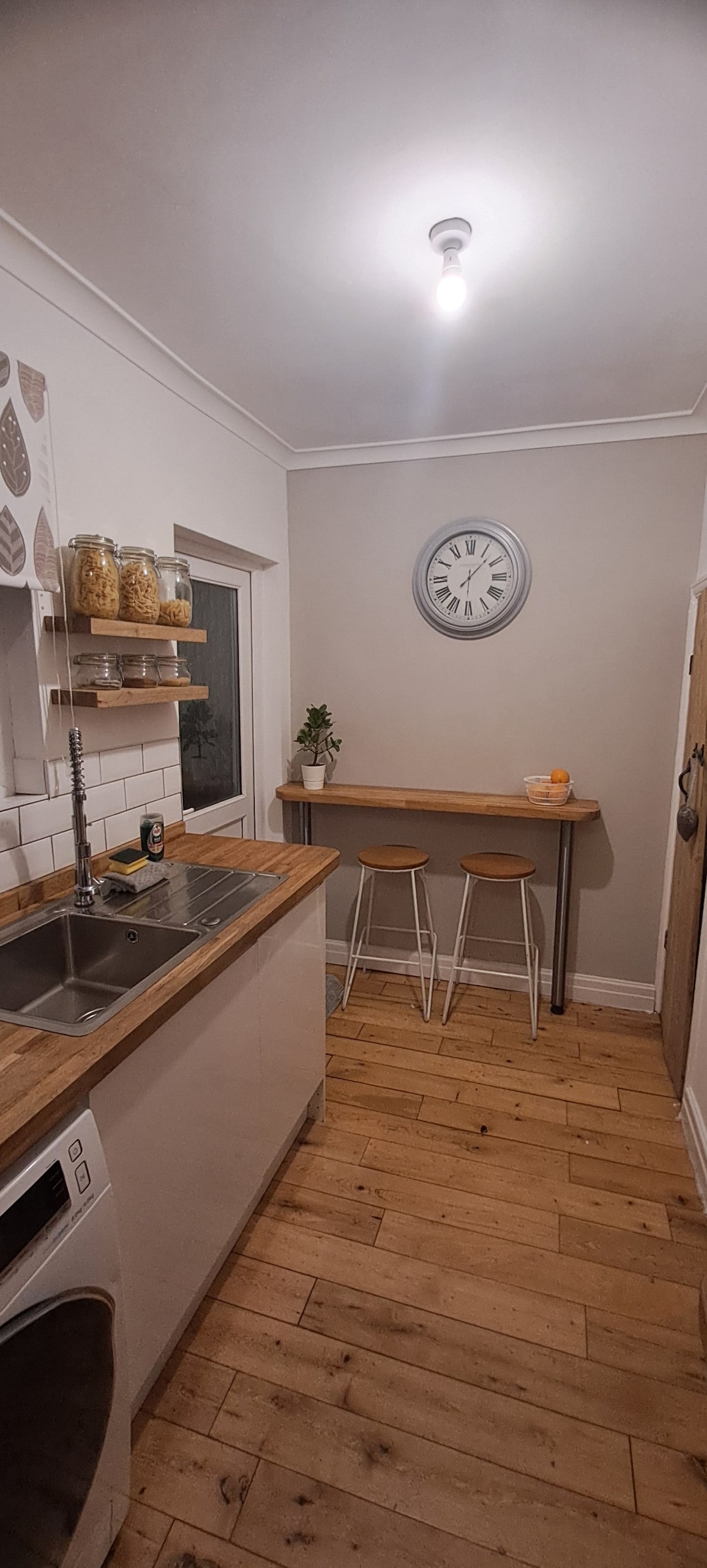 A well-organized kitchen features wooden countertops and light-colored cabinetry. Two wooden stools are positioned at a narrow bar. A wall clock is displayed prominently, while natural light filters through a window. Open shelving holds clear jars, adding a touch of functionality.