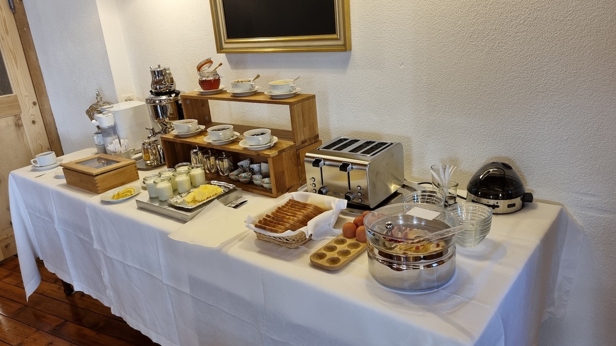A breakfast spread is arranged on a long table covered with a white tablecloth. Various dishes and utensils are displayed, including pastries, eggs, yogurt, and a toaster, creating an inviting setup for guests to enjoy their morning meal.