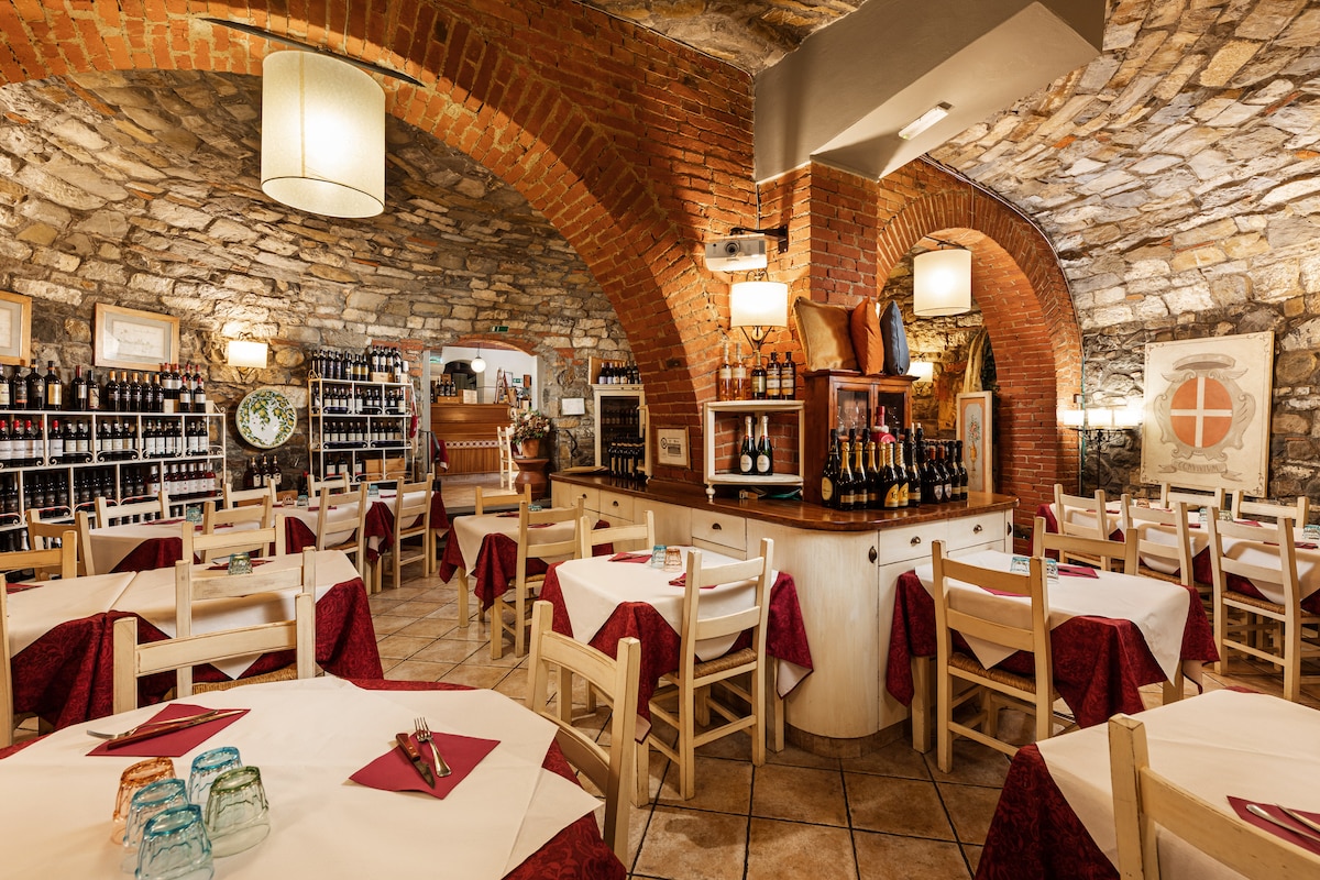 A rustic restaurant interior is featured, characterized by arched, stone walls and warm, ambient lighting. Wooden tables are set with white tablecloths and red napkins. Shelves with assorted wines line the back wall, while large bottles of sparkling wine are prominently displayed on the counter.