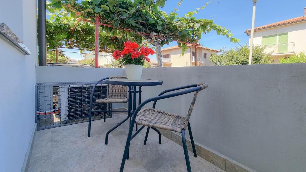 A cozy balcony area is presented with a round table accompanied by two chairs. A potted plant with vibrant red flowers adds a touch of color, while lush greenery overhead creates a shaded atmosphere. The surrounding area features quiet residential buildings.