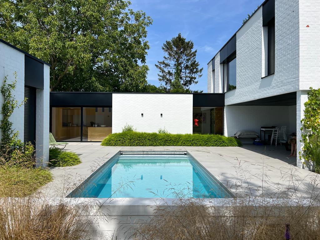 A modern villa is set within a landscaped garden, featuring a heated swimming pool at the center. The structure is framed by greenery, with large windows reflecting the peaceful outdoor space. The patio area offers a serene atmosphere for relaxation.