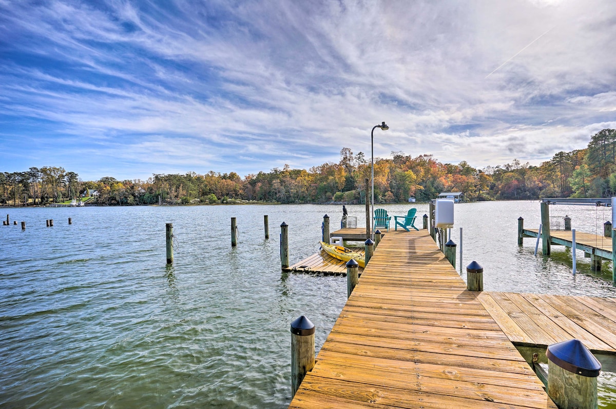 A wooden dock extends into calm waters, surrounded by trees with autumn foliage. Several posts rise from the water, and a pair of turquoise chairs offers a spot for relaxation. A gentle sky with soft clouds creates a serene backdrop.