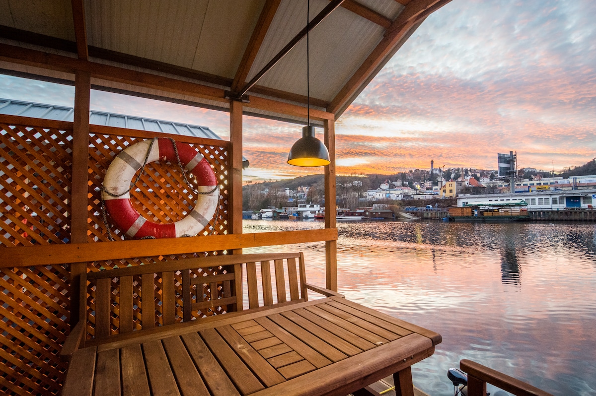 A wooden terrace is shown with a dining table and benches, overlooking a calm waterway. A lifebuoy hangs on the wall nearby, while the sky is filled with colorful clouds at sunset, reflecting in the water.