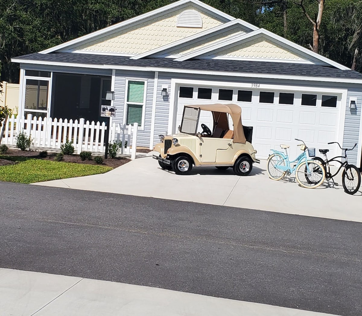 A charming villa exterior is shown, featuring a driveway with a golf cart parked beside two bicycles. Landscaping includes a white picket fence and neatly maintained shrubs. The garage door is visible, with the light-colored home showcasing a classic architectural style.