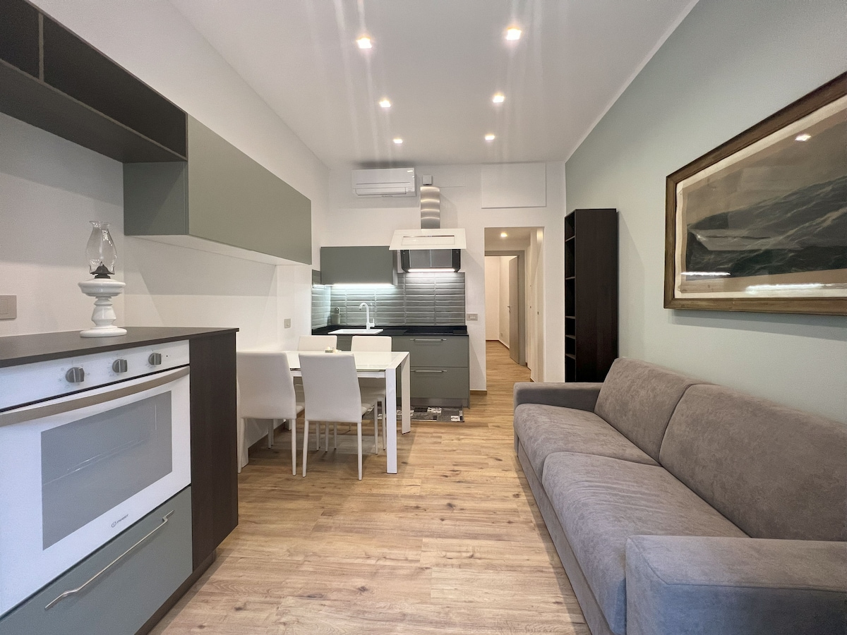 A modern open-concept living area features a sleek kitchen with minimalist cabinetry and a comfortably arranged dining table. A cozy sofa provides seating, and bright overhead lights enhance the space. Neutral tones are complemented by a large wall art piece.