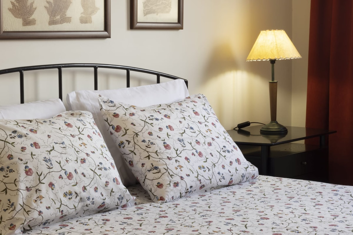 A well-appointed sleeping area features a black metal bed frame with floral-patterned bedding and two decorative pillows. A bedside table holds a gently illuminated lamp, while warm curtains frame the window, adding a touch of comfort to the space.