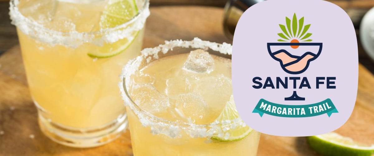 Two glasses filled with a light yellow margarita are displayed on a wooden surface. Each glass is rimmed with salt and garnished with lime wedges. The vibrant logo of the Santa Fe Margarita Trail is featured in the corner.