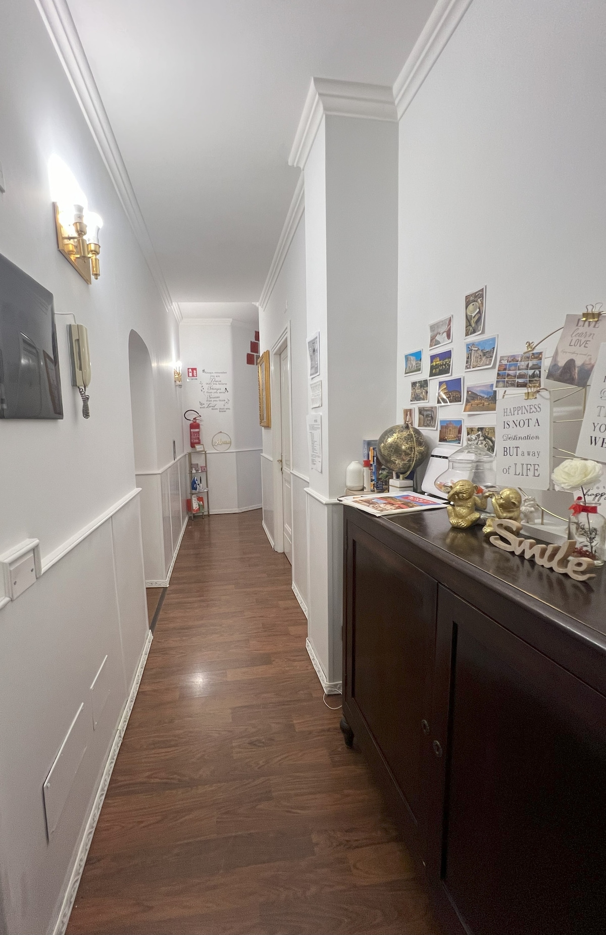 A long corridor is shown, featuring polished wooden flooring and white walls adorned with various framed images and decorative signs. A dark wood cabinet is positioned along the right side, complementing the warm lighting from wall-mounted fixtures.