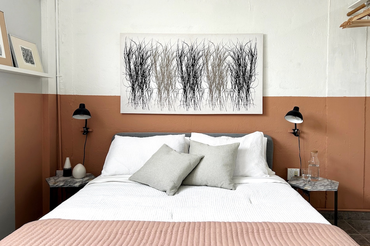 A comfortable bedroom features a spacious bed topped with soft pillows and a textured blanket. Flanking the bed are two bedside lamps placed on marble-topped tables. A large piece of wall art, depicting abstract grass-like forms, adorns the wall above.