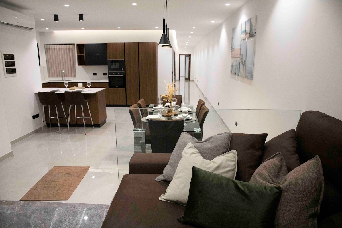 A spacious living area features a comfortable sectional sofa adorned with neutral-toned pillows. A dining table set for four is positioned nearby, with modern kitchen appliances visible in the background. Soft lighting gives a warm ambiance to the open layout.