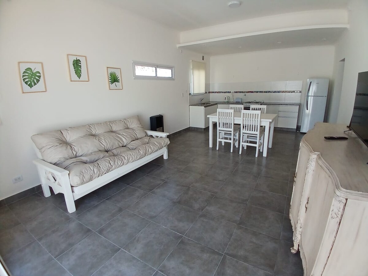A spacious living area features a light-colored futón, white dining table with matching chairs, and a simple cabinet. The kitchen area is visible in the background, showcasing white cabinetry and a refrigerator. Natural light enters through a window, highlighting the neutral tile flooring.