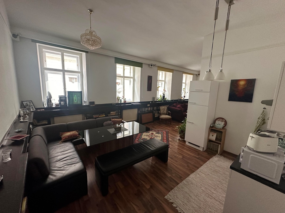 The spacious living area is well-lit by natural light filtering through large windows. A dark, polished table is surrounded by a leather sofa and a long bench. Decorative plants and artwork add character to the room, creating a comfortable gathering space.