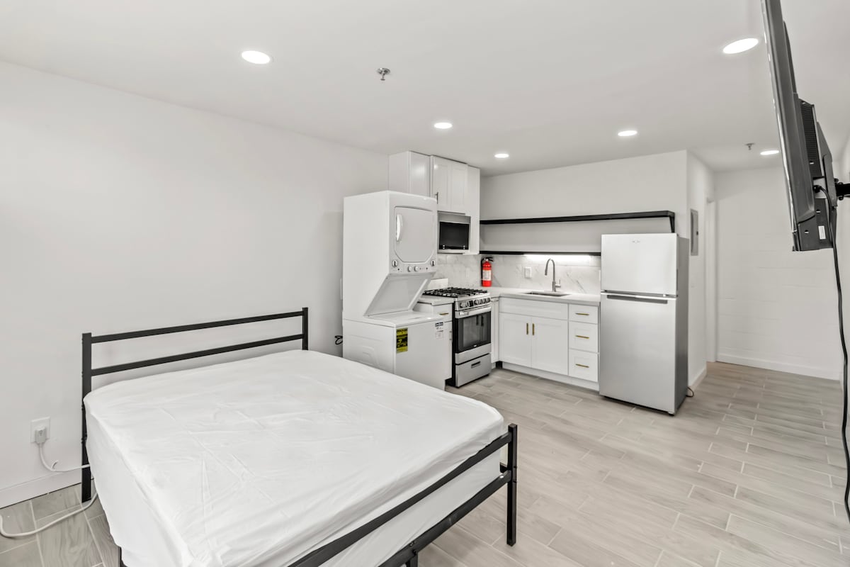 A modern studio apartment is showcased with a minimalist design and sleek finishes. The space includes a metal frame bed, a compact kitchenette with stainless steel appliances, and an open layout that emphasizes the contemporary aesthetic. Bright lighting enhances the overall ambiance.