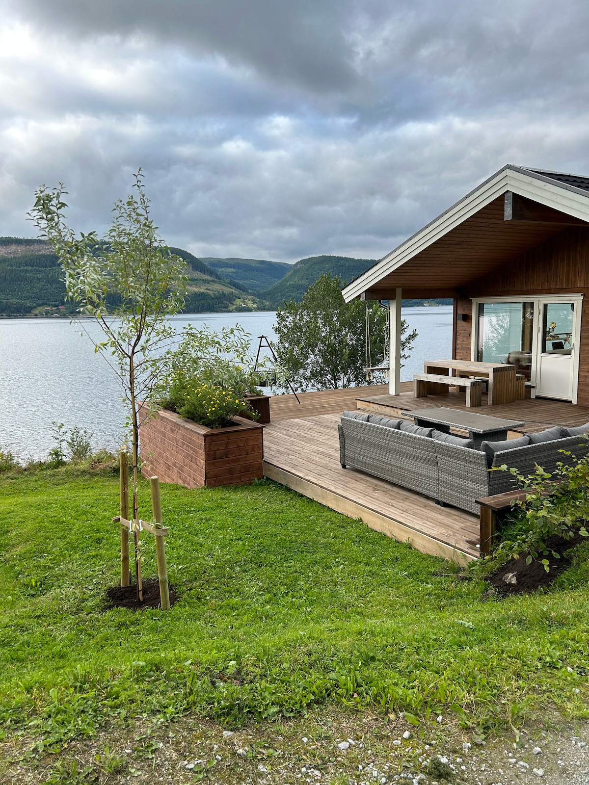 An outdoor deck area features comfortable seating and a dining table, surrounded by greenery and planters. Beyond the deck, a scenic view of the water and mountains is visible, enhancing the serene environment.