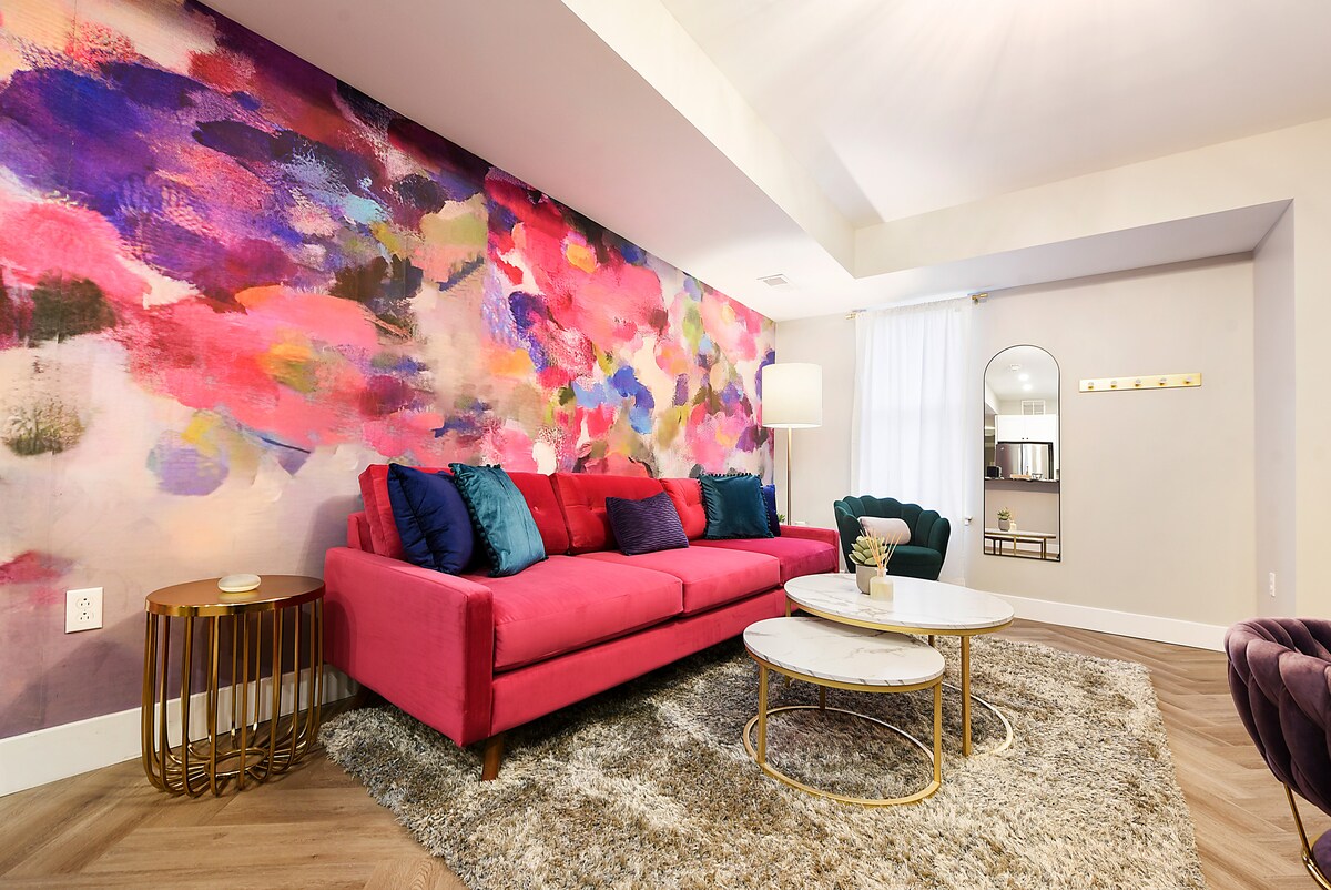 A vibrant living area is presented, featuring a pink Joybird couch complemented by colorful throw pillows. A striking abstract mural decorates the wall behind, while a stylish gold coffee table and a green accent chair add to the modern aesthetic. Natural light filters through the white curtains.
