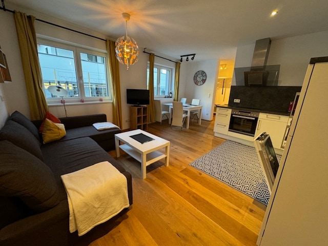 A modern living area is displayed, featuring a spacious dark couch and a white coffee table. Natural light enters through multiple windows, while a dining area with four chairs is visible in the background. A well-equipped kitchen with sleek appliances is situated to the right.