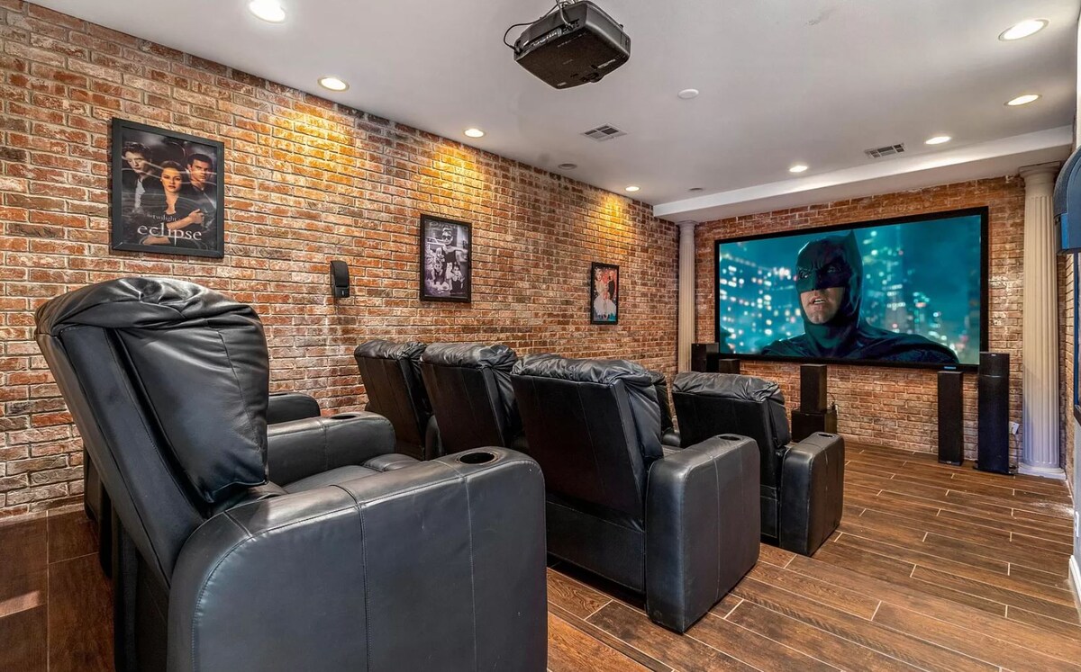 A home theatre room is shown, featuring plush black reclining seats arranged in rows facing a large screen. Exposed brick walls are highlighted by soft lighting, and posters adorn the walls, adding character to the space.