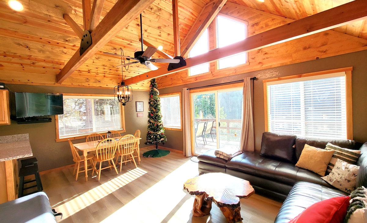 The spacious living area features a high wooden ceiling with exposed beams. Natural light floods in through multiple windows, illuminating a large sectional sofa and a round dining table with several chairs. A small Christmas tree decorates one corner, adding a festive touch.