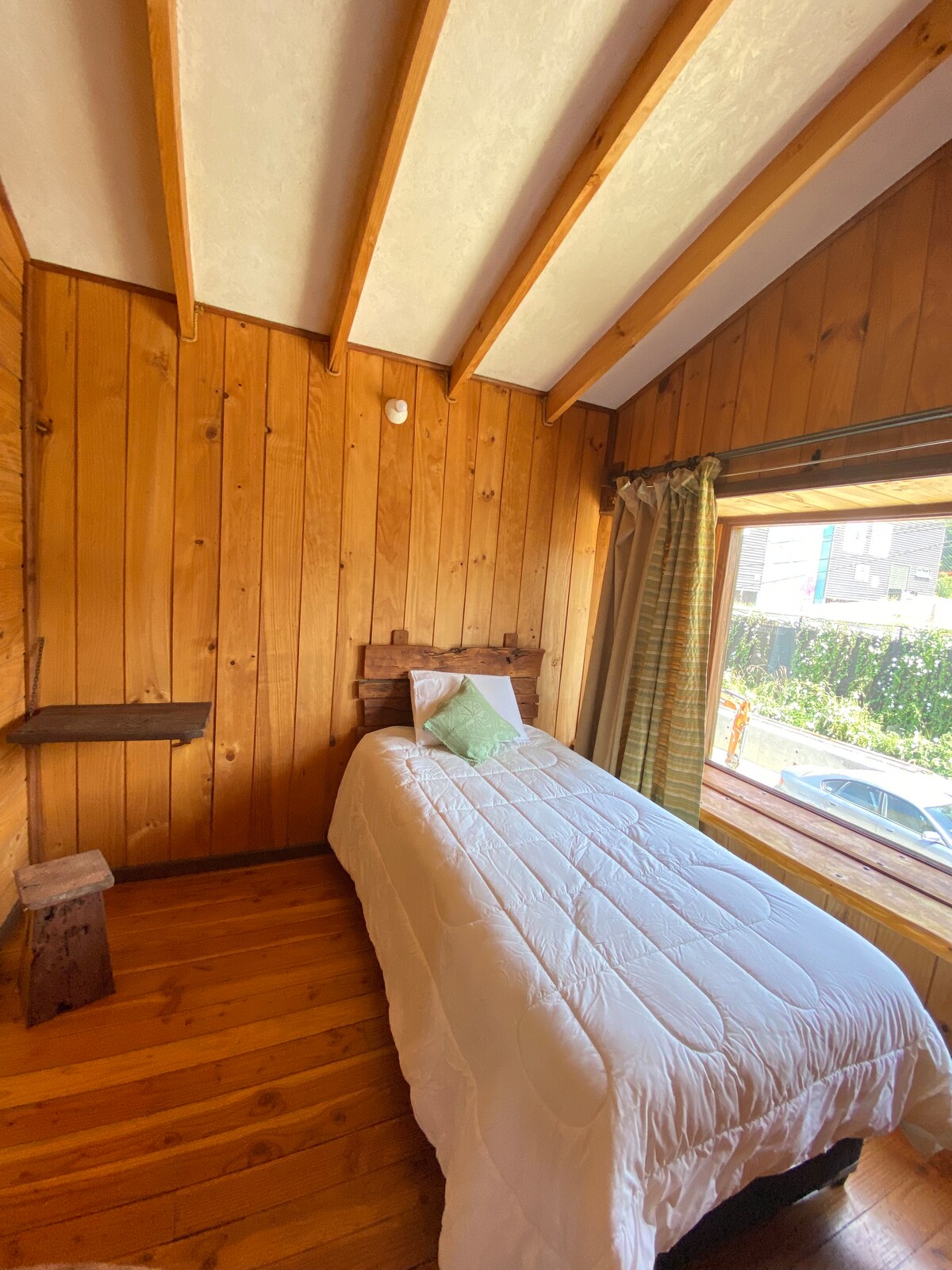 A cozy bedroom features wooden paneling and a sloped ceiling. A narrow bed is dressed in white bedding and positioned beside a large window, allowing natural light to fill the space. Simple shelving is located on the wall, adding functionality.