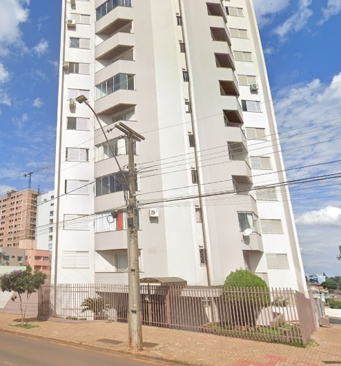 A modern residential building is presented, featuring a sleek white façade. Multiple balconies are visible along the upper levels, while greenery is seen at the ground level, complementing the urban surroundings. The entrance is secured with a gate, promoting a feeling of safety.