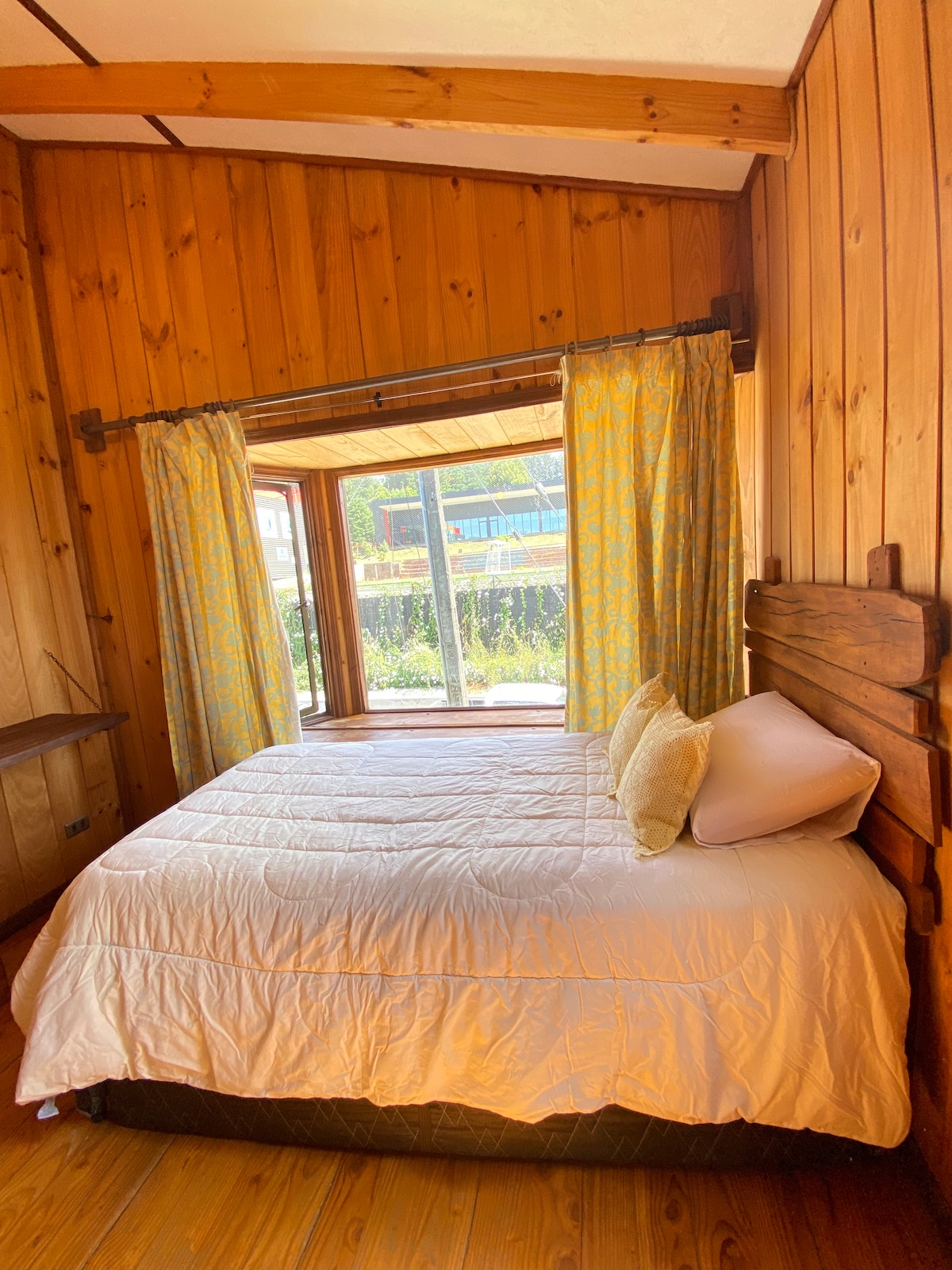 A room features wooden walls and a spacious bed covered with a white quilt. Large windows adorned with light yellow curtains allow natural light to enter, providing views of the surrounding greenery.