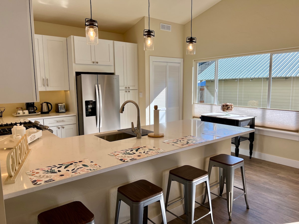 A modern kitchen features a spacious island with four metal stools positioned around it. Pendant lights hang above, illuminating the countertop. A large window provides natural light, overlooking a yard with a green roof building visible outside.