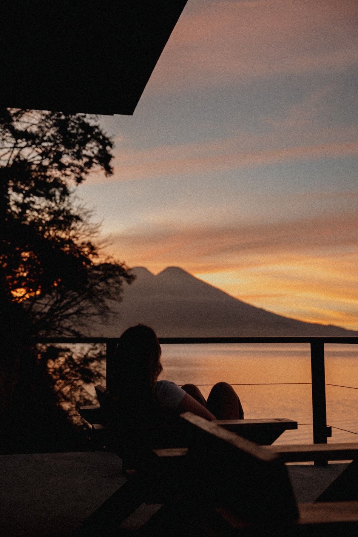 A peaceful sunset view is captured from a balcony, featuring silhouettes of two volcanoes against a colorful sky. A person is seated on a wooden lounge chair, gazing at the tranquil waters below, surrounded by trees on the hillside.