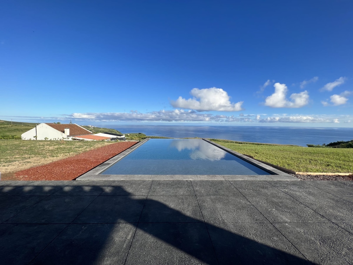 An infinity edge saltwater pool reflects the open sky and ocean vistas, creating a serene ambiance. The surrounding grassy area offers space for relaxation, while the distant coastline is visible under expansive blue skies.