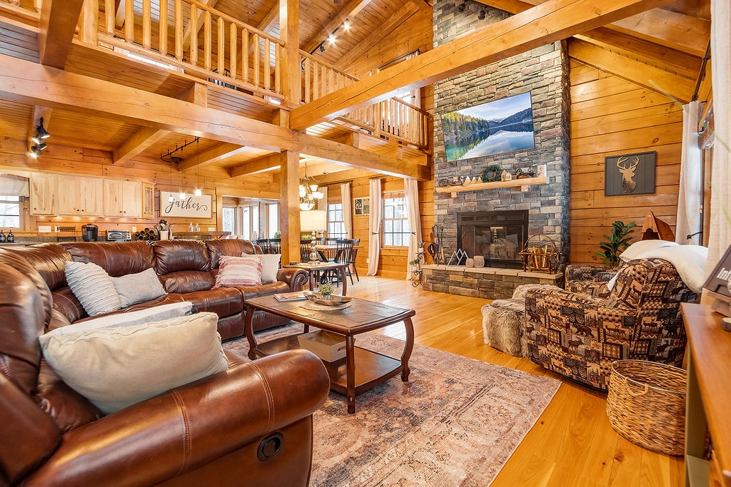 The open-concept living area features a wood-burning fireplace surrounded by stone, with warm wooden beams overhead. Cozy leather sofas are arranged around a wooden coffee table, while natural light flows through large windows, illuminating the rustic decor and inviting atmosphere.