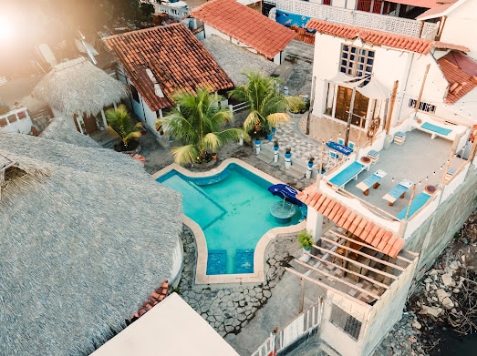 An aerial view captures the layout of multiple bungalows surrounding a spacious swimming pool. Palms provide shade and color. Sun loungers are arranged near the pool, complemented by wooden structures and ceramic tiles in the outdoor areas, creating a harmonious outdoor environment.