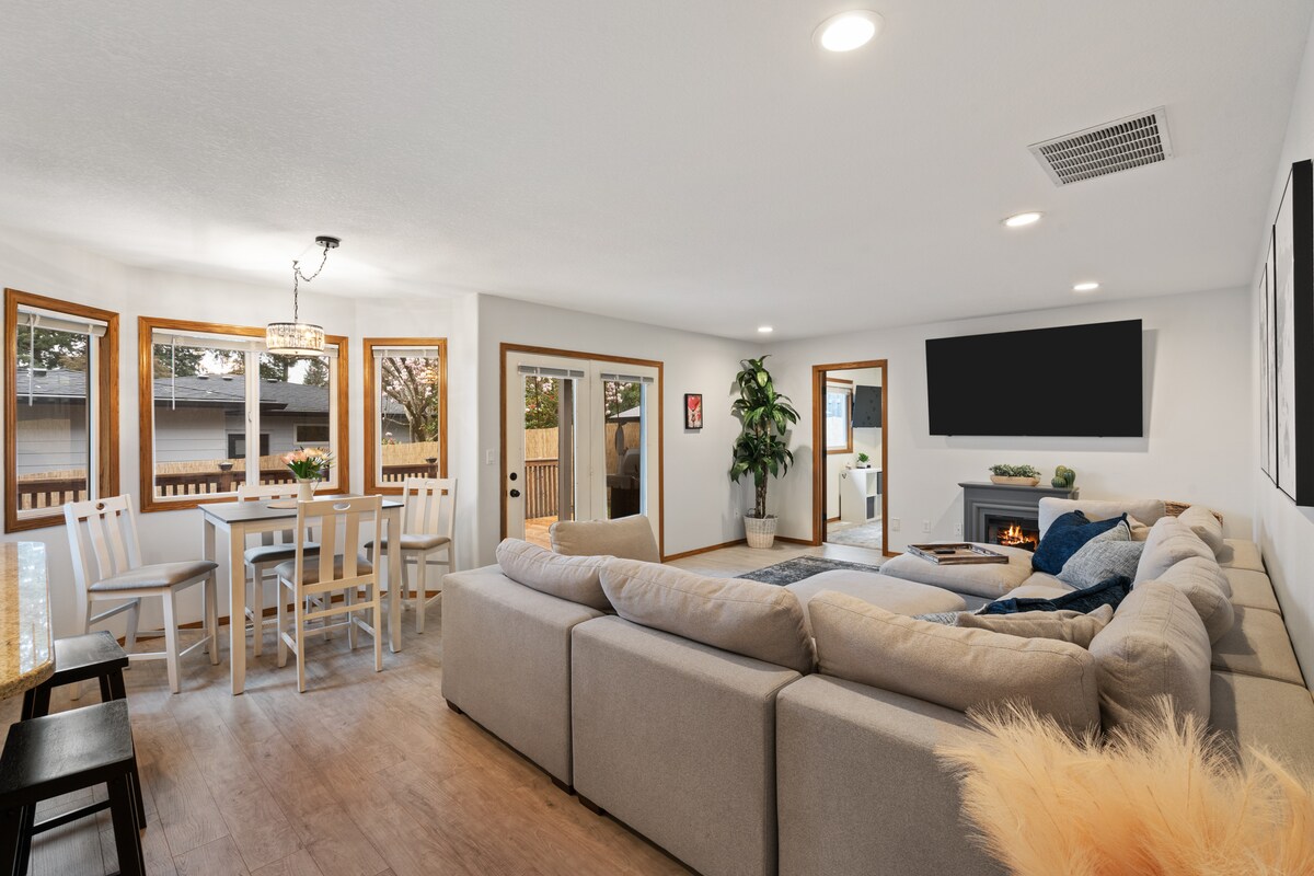 A spacious living area features a large sectional sofa in light neutral tones. Natural light fills the room through multiple windows. A modern dining table is positioned nearby, complemented by a small indoor plant. A flat-screen television is mounted on the wall above an electric fireplace.
