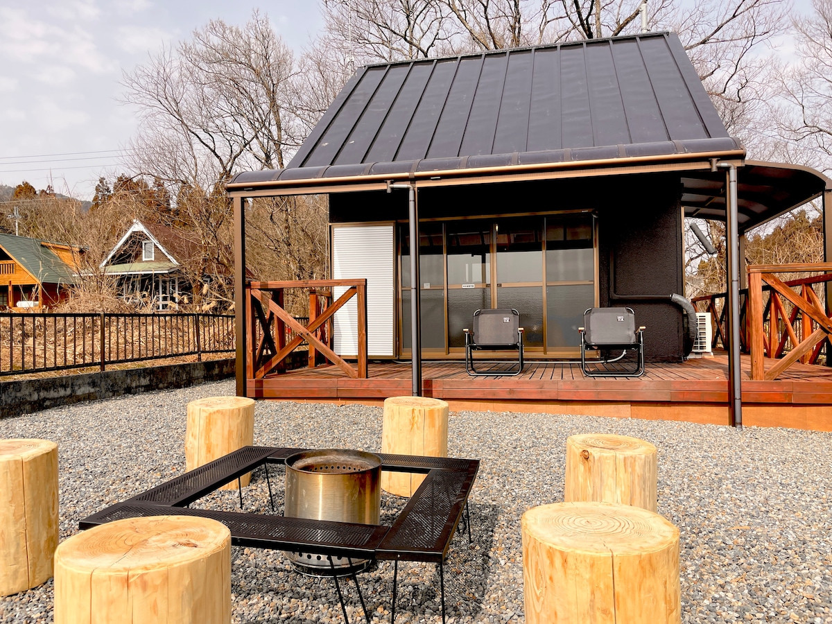 A spacious outdoor area features a wooden deck connected to a cottage. Natural log stools are arranged around a fire pit, with a gravel surface extending towards the surrounding landscape. The structure is fitted with large windows, providing views of the garden.