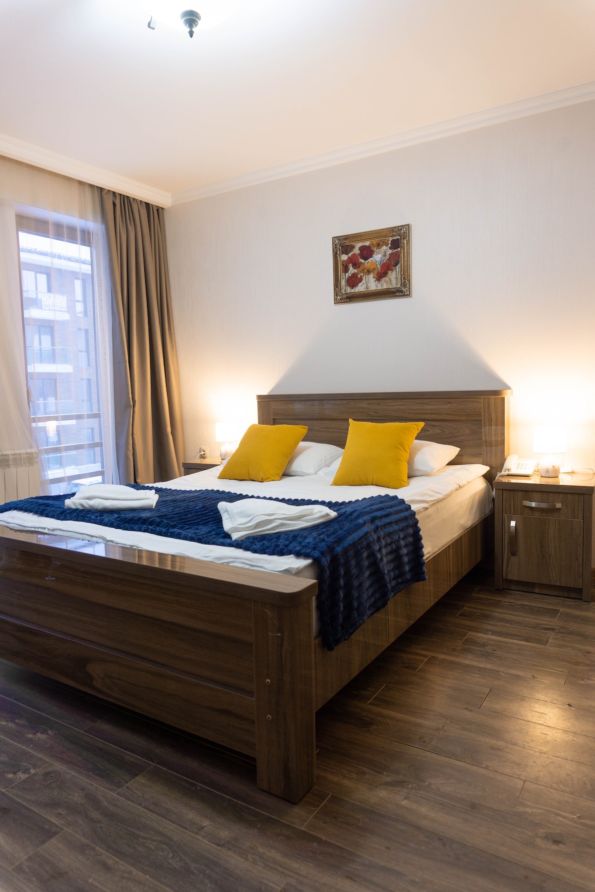 A spacious bedroom is furnished with a wooden bed featuring a lavish 2-person mattress, adorned with blue and white bedding. Two yellow pillows add a touch of color. Soft lighting is provided by bedside lamps, and natural light filters through the curtains of the window.
