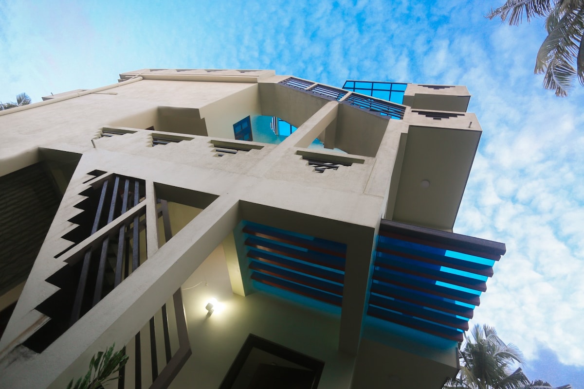 The exterior of a multi-level building is captured from a low angle, showcasing the architectural details and modern design elements. The facade features a mix of horizontal and vertical lines, complemented by large windows and blue accents against a backdrop of a bright sky with scattered clouds.