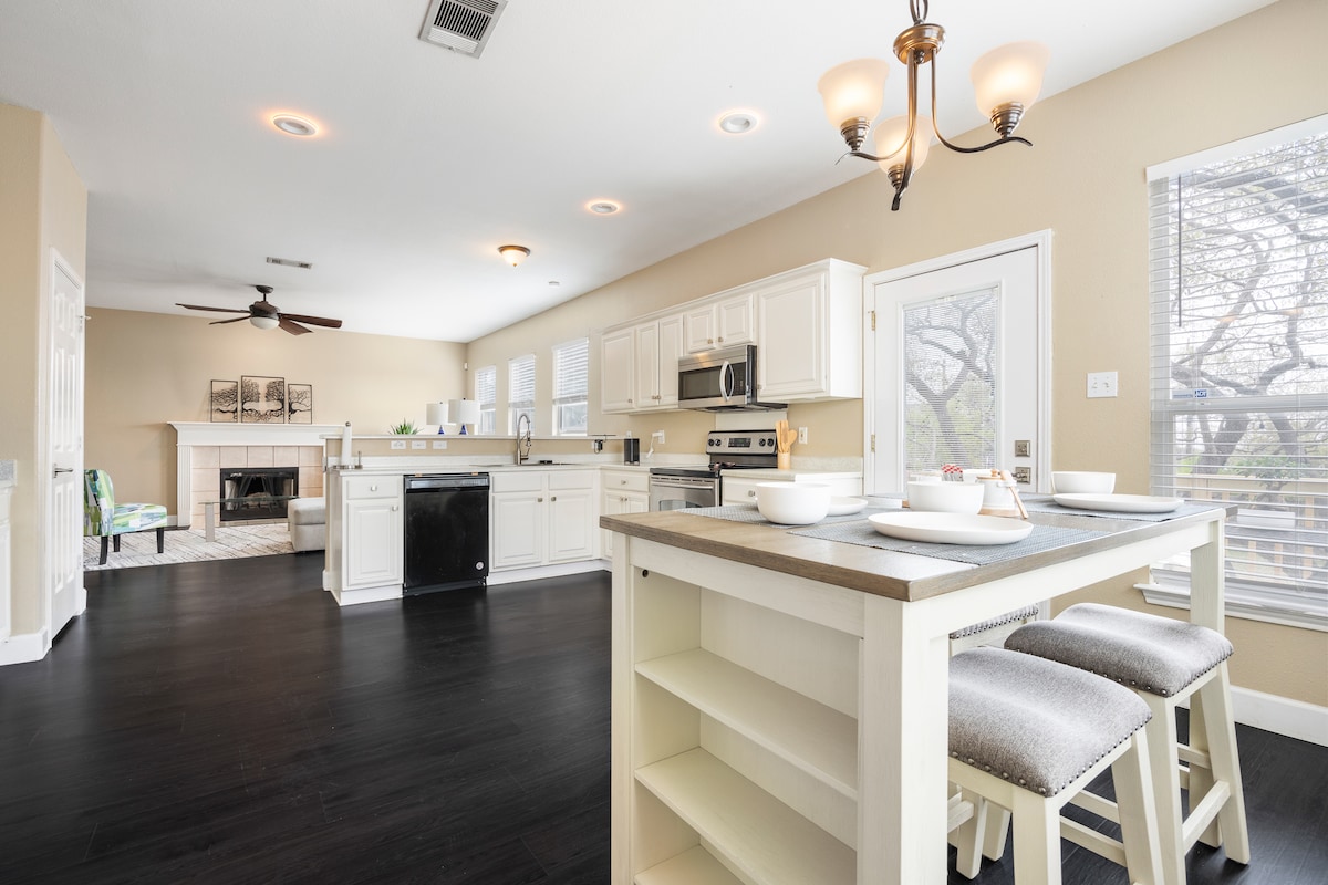 An open-concept kitchen and dining area features white cabinetry and a central island with seating for three. Natural light enters through large windows, illuminating the space. A cozy fireplace is visible in the living area, creating a warm atmosphere.